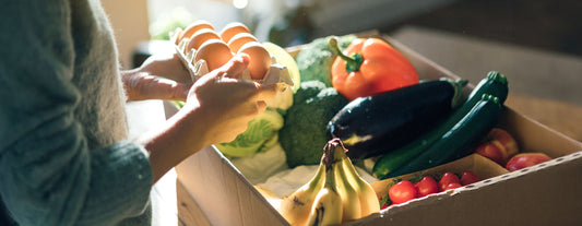 A menopausal woman is holding a carton of eggs. In the background, there is a box of fresh fruits and vegetables to help boost menopause health.