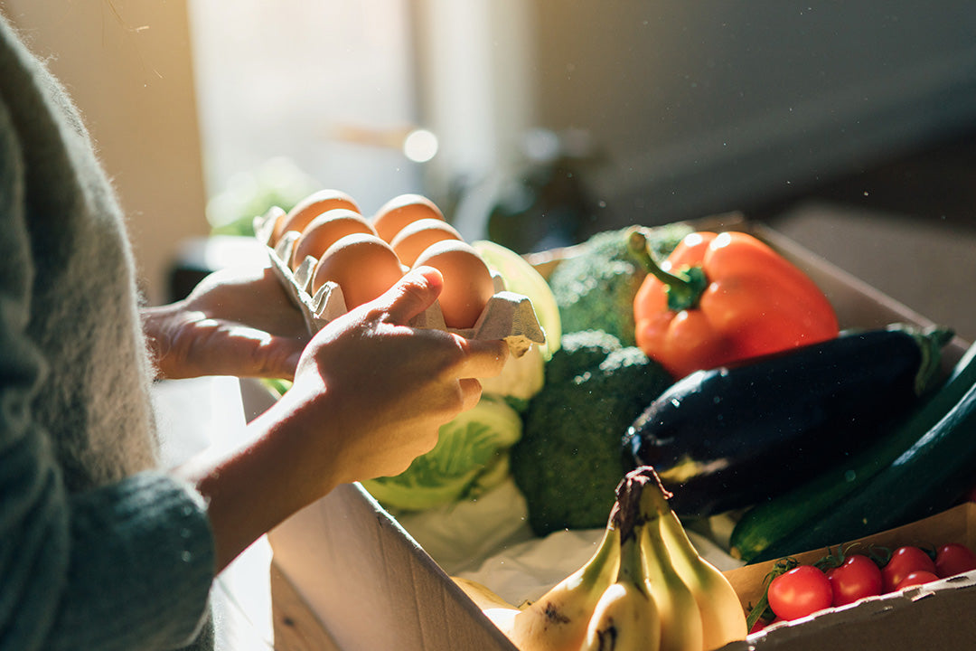 A menopausal woman is holding a carton of eggs. In the background, there is a box of fresh fruits and vegetables to help boost menopause health.